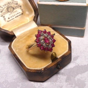 Op de afbeelding: Een gouden ring met een cluster van rode robijnen en witte diamanten, gezet in een stervormig ontwerp. De ring bevindt zich in een kleine houten doos met een fluwelen voering.