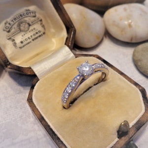 Op de afbeelding: Een diamanten verlovingsring met een enkele ronde briljant gestoken in een witgouden band. De ring zit in een bruine fluwelen ringdoos met de inscriptie "G.F. Truscott, Opticien, Bideford, Telefoon Bideford 117".