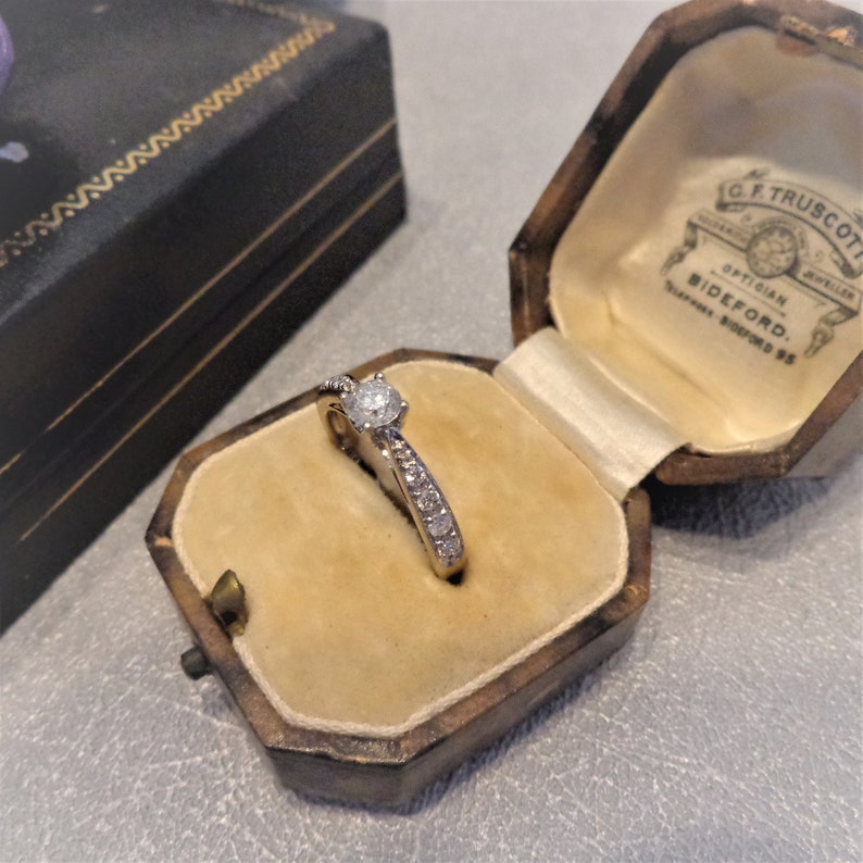 Op de afbeelding: Een diamanten verlovingsring met een enkele ronde steen en kleinere stenen die in een band zijn gezet. De ring bevindt zich in een bruine fluwelen ringdoos met de inscriptie "G.F. Truscott Optician Jeweller Bideford."