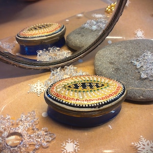 May include: A small, oval-shaped trinket box with a blue base and a white lid. The lid is decorated with a gold and white floral design and a black eye-like shape in the centre.