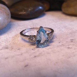 以下が含まれることがあります： 中央に洋梨型の水色の宝石をあしらったシルバーリング。両側に2つの小さなクリアストーンが配置されています。リングは、テクスチャ加工されたオフホワイトの表面に置かれ、背景はぼやけています。