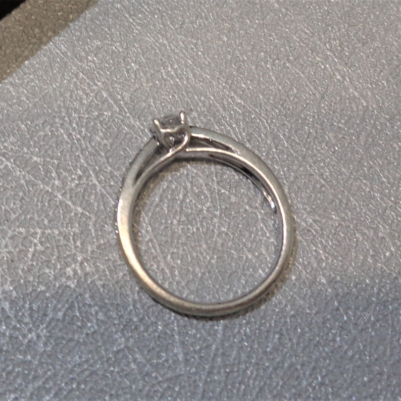 Op de afbeelding: Een zilveren ring met een kleine, heldere edelsteen in een kraalbezetting. De band is dun en heeft een rij kleine, heldere edelstenen.