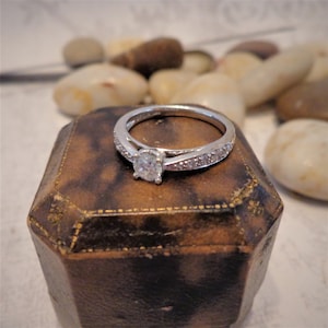 Op de afbeelding: Een zilveren verlovingsring met een enkele ronde diamant en een band met kleinere diamanten. De ring ligt in een bruine fluwelen ringdoos.