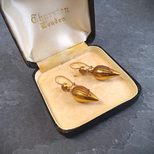 May include: A pair of gold-toned drop earrings with a pointed, teardrop-shaped base and delicate wire-wrapped detailing, presented in a black hinged jewelry box with a cream-colored interior featuring the text Thurston London printed in gold.