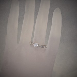 Op de afbeelding: Een zilveren ring met een enkele, ronde, heldere steen in het midden en kleinere heldere stenen die in een band rond de steen zijn gezet.