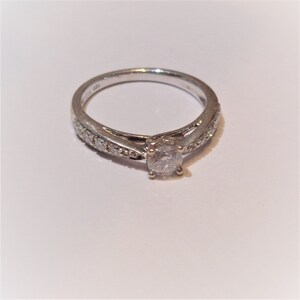 Op de afbeelding: Een zilveren ring met een enkele ronde diamant in het midden en kleinere diamanten die in een band rond de ring zijn gezet.