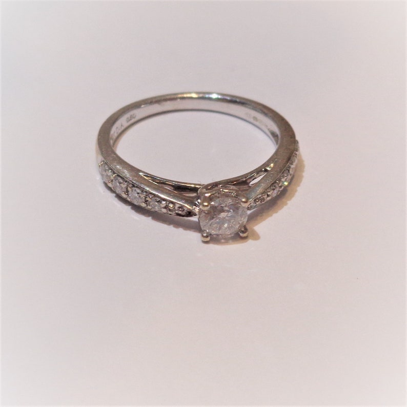 Op de afbeelding: Een zilveren ring met een enkele ronde diamant in het midden en kleinere diamanten die in een band rond de ring zijn gezet.