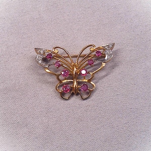 Puede incluir: Broche de mariposa dorada con gemas de color rubí y detalles de diamantes. Las alas de la mariposa están abiertas, y el broche está sobre un fondo gris claro. Una delicada pieza de joyería.
