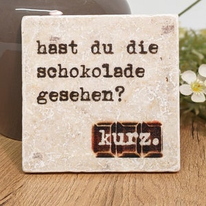 Puede incluir: Un posavasos cuadrado, blanquecino, con el texto alemán "hast du die schokolade gesehen?" y la palabra "kurz." en un rectángulo marrón. El posavasos tiene una superficie texturizada y una estética vintage.