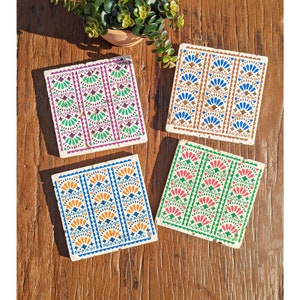 Può includere: Quattro sottobicchieri quadrati con motivi colorati e ripetuti. Ogni sottobicchiere presenta una combinazione di colori diversa: viola e verde, marrone e blu, blu e arancione e rosso e verde. I sottobicchieri sono su una superficie di legno, con una piccola pianta in vaso sullo sfondo.