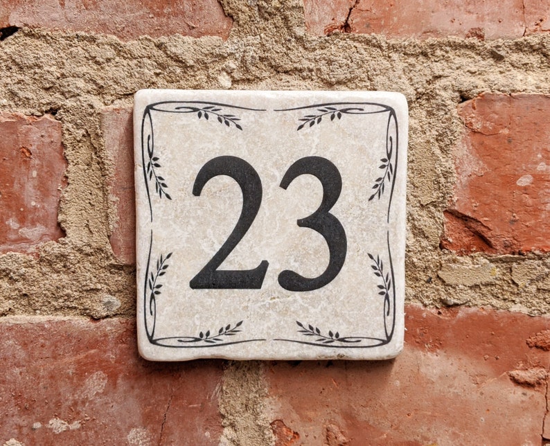 Puede incluir: Placa de n&uacute;mero de casa cuadrada de cer&aacute;mica blanquecina con el n&uacute;mero 23 en negro. La placa est&aacute; decorada con un borde floral negro y est&aacute; montada en una pared de ladrillo rojo.