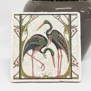 Può includere: Sottobicchiere quadrato in ceramica con un'illustrazione stilizzata di due aironi. Gli uccelli hanno piumaggio nero e verde, becchi e zampe arancioni. Il design è incorniciato da un bordo decorativo con accenti rossi.