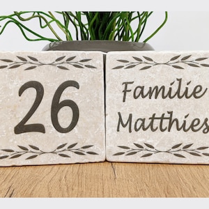 Può includere: Due piastrelle di pietra beige con testo marrone scuro e disegni di foglie. Una piastrella mostra il numero "26", l'altra dice "Familie Matthies". Le piastrelle sono su una superficie di legno, con vegetazione sullo sfondo.