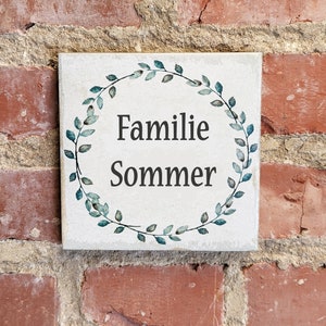 May include: A square, off-white tile with the words "Familie Sommer" in black, encircled by a watercolor-style wreath of blue-green leaves. The tile is mounted on a weathered red brick wall.