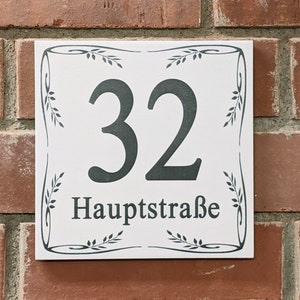 Könnte beinhalten: Quadratisches weißes Keramik-Hausnummernschild mit der Nummer 32 in Dunkelgrau und dem Wort "Hauptstraße" darunter. Das Schild ist mit einem dekorativen Blattmuster umrandet und an einer roten Backsteinmauer befestigt.