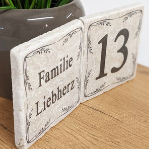 Puede incluir: Dos baldosas de piedra blanquecina con texto negro y bordes decorativos. Una baldosa dice "Familie Liebherz", la otra muestra el n&uacute;mero "13". Las baldosas est&aacute;n colocadas sobre una superficie de madera, con una maceta gris y vegetaci&oacute;n al fondo.