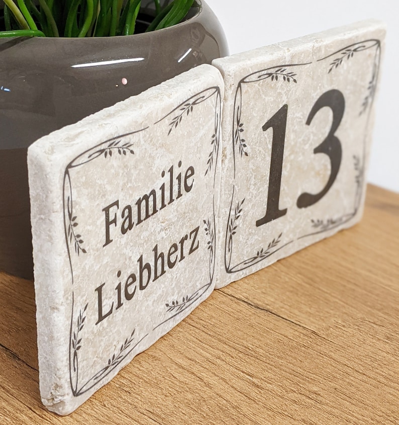 Puede incluir: Dos baldosas de piedra blanquecina con texto negro y bordes decorativos. Una baldosa dice "Familie Liebherz", la otra muestra el n&uacute;mero "13". Las baldosas est&aacute;n colocadas sobre una superficie de madera, con una maceta gris y vegetaci&oacute;n al fondo.