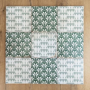 Könnte beinhalten: Neun quadratische Keramikfliesen, die in einem Raster angeordnet sind. Die Fliesen haben einen weißen Hintergrund mit einem sich wiederholenden dunkelgrünen Art-Deco-Fächerdesign und einem kleineren floralen Muster. Die Fliesen werden auf einer hellbraunen Holzoberfläche präsentiert.