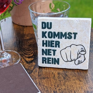 Pode incluir: Uma base de pedra quadrada com o texto alemão "DU KOMMST HIER NET REIN" em preto, e um desenho de uma mão a apontar. Uma taça de vinho tinto, um copo transparente e um pequeno caderno estão sobre a mesa de madeira.