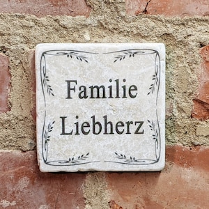 Puede incluir: Placa cuadrada de piedra blanquecina con las palabras "Familie Liebherz" en letras negras. La placa est&aacute; enmarcada con un dise&ntilde;o floral negro y montada sobre una pared de ladrillo rojo.