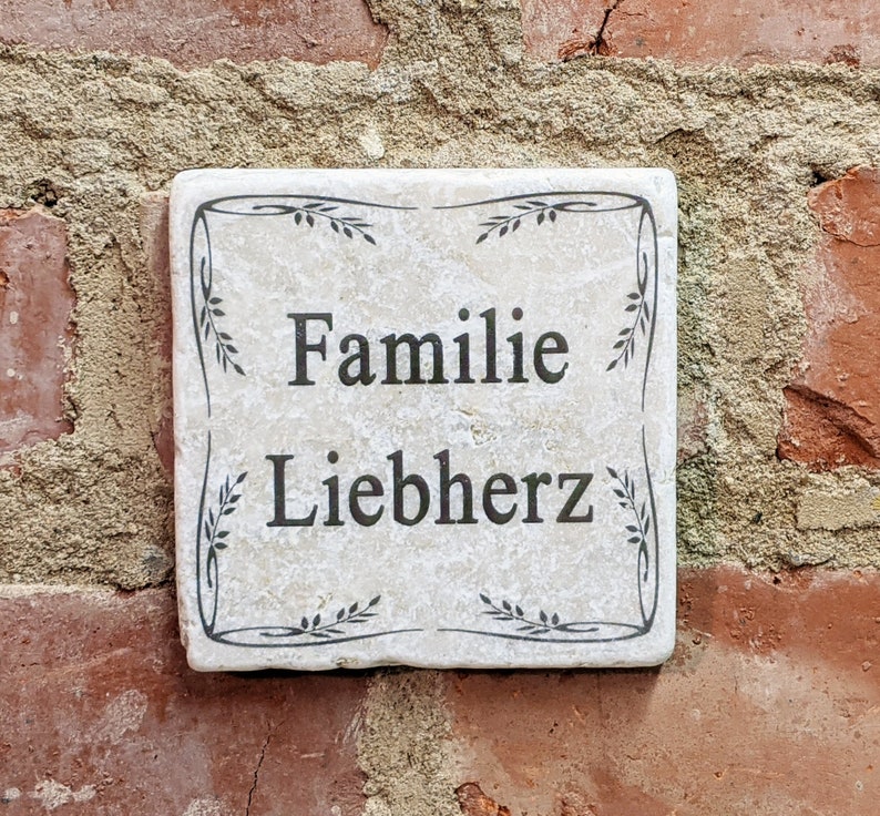 Puede incluir: Placa cuadrada de piedra blanquecina con las palabras "Familie Liebherz" en letras negras. La placa est&aacute; enmarcada con un dise&ntilde;o floral negro y montada sobre una pared de ladrillo rojo.