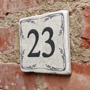 Puede incluir: Placa de n&uacute;mero de casa cuadrada de cer&aacute;mica con el n&uacute;mero 23 en negro. La placa tiene un fondo crema con un borde floral negro. Est&aacute; montada en una pared de ladrillo rojo.