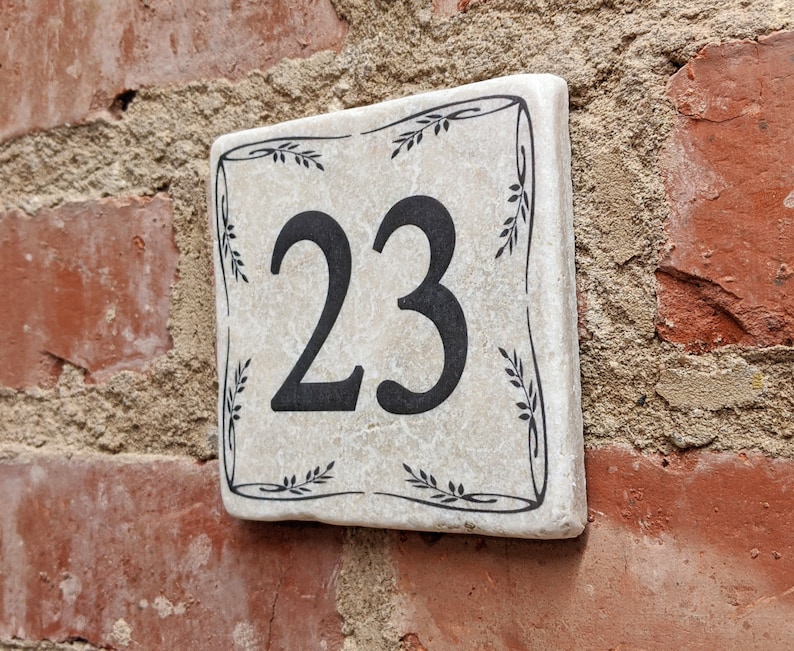 Puede incluir: Placa de n&uacute;mero de casa cuadrada de cer&aacute;mica con el n&uacute;mero 23 en negro. La placa tiene un fondo crema con un borde floral negro. Est&aacute; montada en una pared de ladrillo rojo.