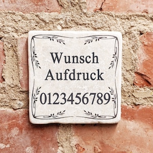 Puede incluir: Una baldosa cuadrada de cer&aacute;mica blanquecina con texto negro y un borde decorativo de hojas. El texto dice "Wunsch Aufdruck" y una serie de n&uacute;meros. La baldosa est&aacute; montada en una pared de ladrillo rojo.