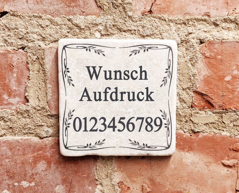 Puede incluir: Una baldosa cuadrada de cer&aacute;mica blanquecina con texto negro y un borde decorativo de hojas. El texto dice "Wunsch Aufdruck" y una serie de n&uacute;meros. La baldosa est&aacute; montada en una pared de ladrillo rojo.