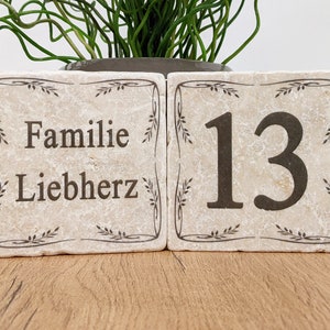 Puede incluir: Dos bloques de piedra beige con texto marr&oacute;n oscuro y bordes decorativos. Un bloque dice "Familie Liebherz", el otro muestra el n&uacute;mero "13". Los bloques est&aacute;n sobre una superficie de madera, con una planta en el fondo.