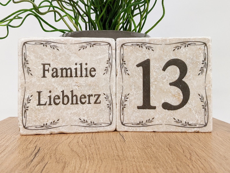 Puede incluir: Dos bloques de piedra beige con texto marr&oacute;n oscuro y bordes decorativos. Un bloque dice "Familie Liebherz", el otro muestra el n&uacute;mero "13". Los bloques est&aacute;n sobre una superficie de madera, con una planta en el fondo.