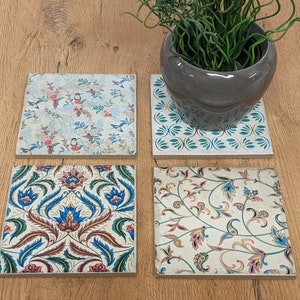 Op de afbeelding: Vier vierkante keramische onderzetters met bloempatronen. Eén heeft een turquoise en wit ontwerp, andere hebben ingewikkelde bloemen- en vogelmotieven in blauw, rood en groen. Een grijze pot met een groene plant staat op een onderzetter. Ongeveer 10 cm x 10 cm.