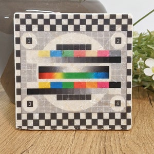 Könnte beinhalten: Quadratischer Keramikuntersetzer mit Retro-TV-Testmuster-Design. Der Untersetzer hat einen schwarz-weißen Schachbrettmuster-Rand und eine zentrale Grafik mit farbigen horizontalen Balken und nummerierten Kreisen.