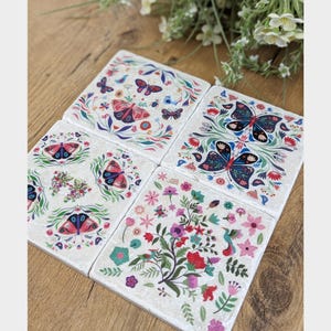 Könnte beinhalten: Vier quadratische Untersetzer mit Schmetterlings- und Blumendesigns. Jeder Untersetzer zeigt eine einzigartige Anordnung von bunten Schmetterlingen, Blumen und Blättern. Die Untersetzer sind weiß mit einer leicht strukturierten Oberfläche.