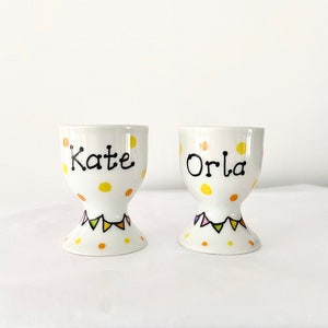 May include: Two white ceramic egg cups with yellow polka dots and colourful bunting around the base. The cups are personalised with the names "Kate" and "Orla" in black.