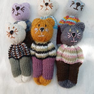 May include: A collection of six small, hand-knitted animal dolls with round heads and embroidered facial features. Each doll wears a unique jumper with varied patterns and colours, including stripes and solid tones, paired with matching knitted trousers.