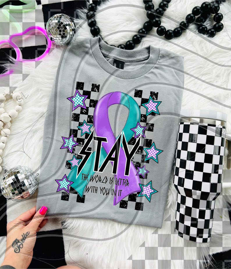 May include: A gray t-shirt with a purple and teal ribbon design and the word "STAY". The shirt also features a checkered pattern and star accents. A black and white checkered tumbler is also visible.