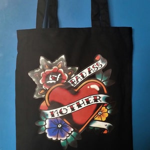 May include: Black tote bag with a heart-shaped tattoo design. The heart is red with a banner that reads "BAD ASS MOTHER". The design includes flowers and leaves. The bag has black handles.