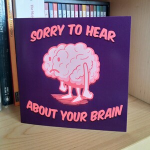 May include: A purple card with a pink cartoon brain with a sad expression. The text "Sorry to hear about your brain" is printed in pink.