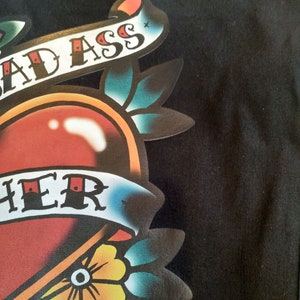 May include: Close-up of a black garment featuring a colorful tattoo-style graphic. The design includes a red heart, a banner with the words "BAD ASS" and "HER", and floral elements in yellow, blue, and green.