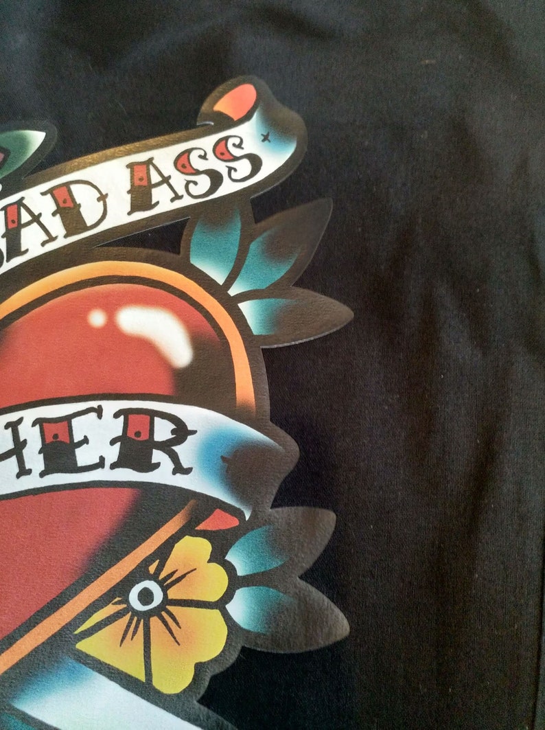 May include: Close-up of a black garment featuring a colorful tattoo-style graphic. The design includes a red heart, a banner with the words "BAD ASS" and "HER", and floral elements in yellow, blue, and green.