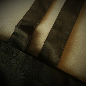 May include: Close-up of a dark green tote bag with two matching straps. The straps are sewn onto the bag with visible stitching. The background is a light beige fabric.