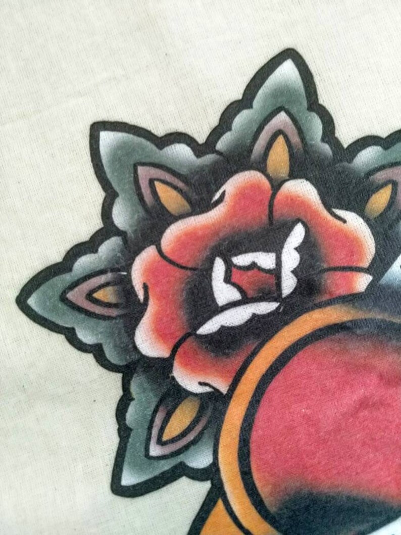 May include: A close-up of a traditional tattoo design featuring a red rose with green and black leaves. The rose is surrounded by a yellow and orange circle.