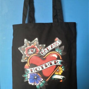 May include: Black tote bag with a tattoo-style graphic. The design features a red heart with a banner reading "MOTHER" and "BAD ASS" on a banner. Floral elements surround the heart, with a blue background.