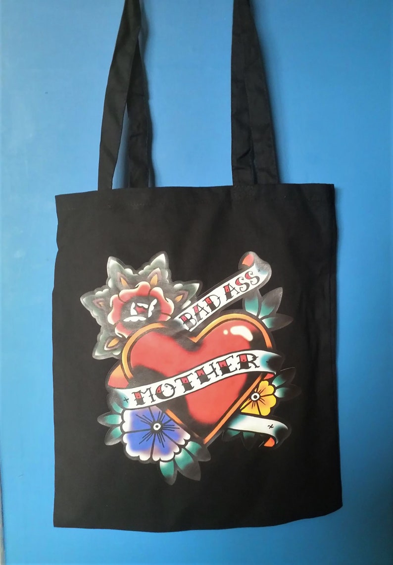 May include: Black tote bag with a tattoo-style graphic. The design features a red heart with a banner reading "MOTHER" and "BAD ASS" on a banner. Floral elements surround the heart, with a blue background.
