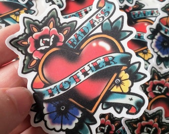 May include: A white sticker with a red heart, a banner that says "Mother" and another banner that says "Badass". The heart is surrounded by flowers in shades of red, blue, yellow, and green.