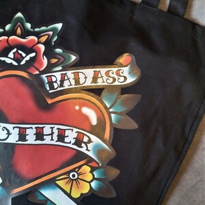May include: Black tote bag featuring a heart-shaped tattoo design with a banner that reads "BAD ASS MOTHER". The design includes a red heart, a rose, and other floral elements in a classic tattoo style.