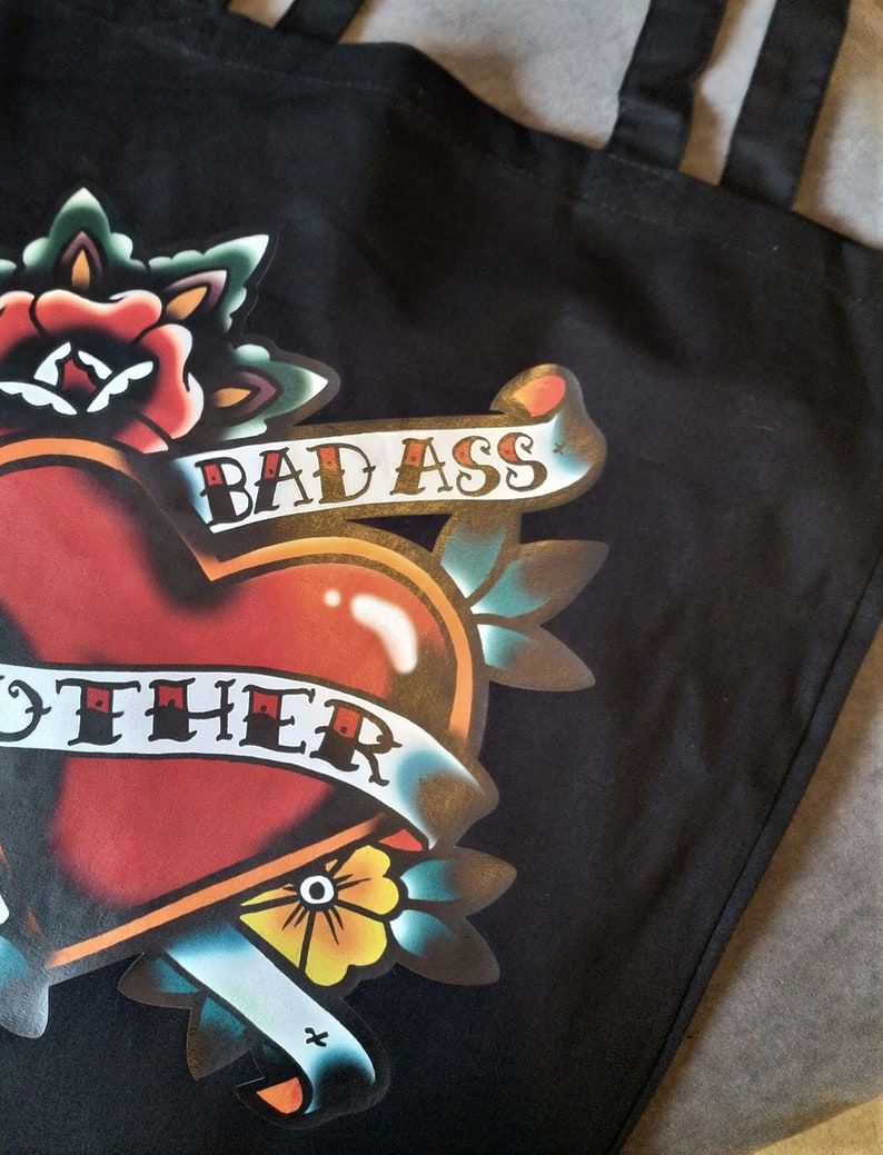 May include: Black tote bag featuring a heart-shaped tattoo design with a banner that reads "BAD ASS MOTHER". The design includes a red heart, a rose, and other floral elements in a classic tattoo style.