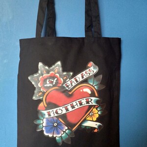 May include: Black tote bag with a tattoo-style graphic. The design features a red heart with a banner that reads "MOTHER" and "BAD ASS". The graphic includes floral and leaf elements in various colors.