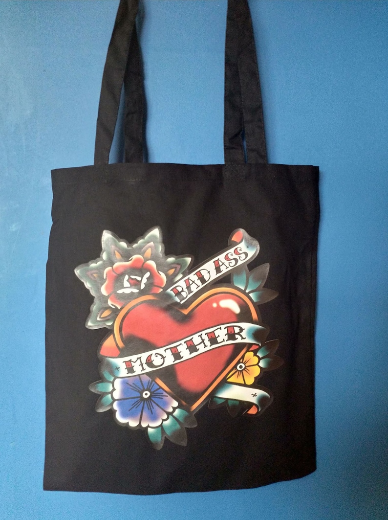 May include: Black tote bag with a tattoo-style graphic. The design features a red heart with a banner that reads "MOTHER" and "BAD ASS". The graphic includes floral and leaf elements in various colors.